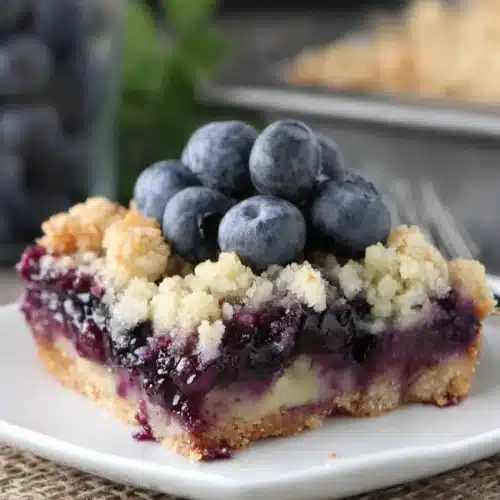 Delicious Blueberry Pie Bars You’ll Love 1 A slice of blueberry pie bar on a white plate with fresh blueberries