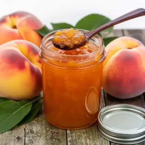 Low Sugar Peach Jam: Easy & Delicious Recipe 2 Low sugar peach jam in a glass jar with fresh peaches and green leaves on a wooden table