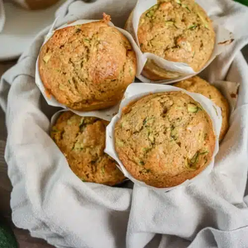 Sourdough discard zucchini muffins in white liners on a cloth, fresh and moist with visible zucchini and walnuts